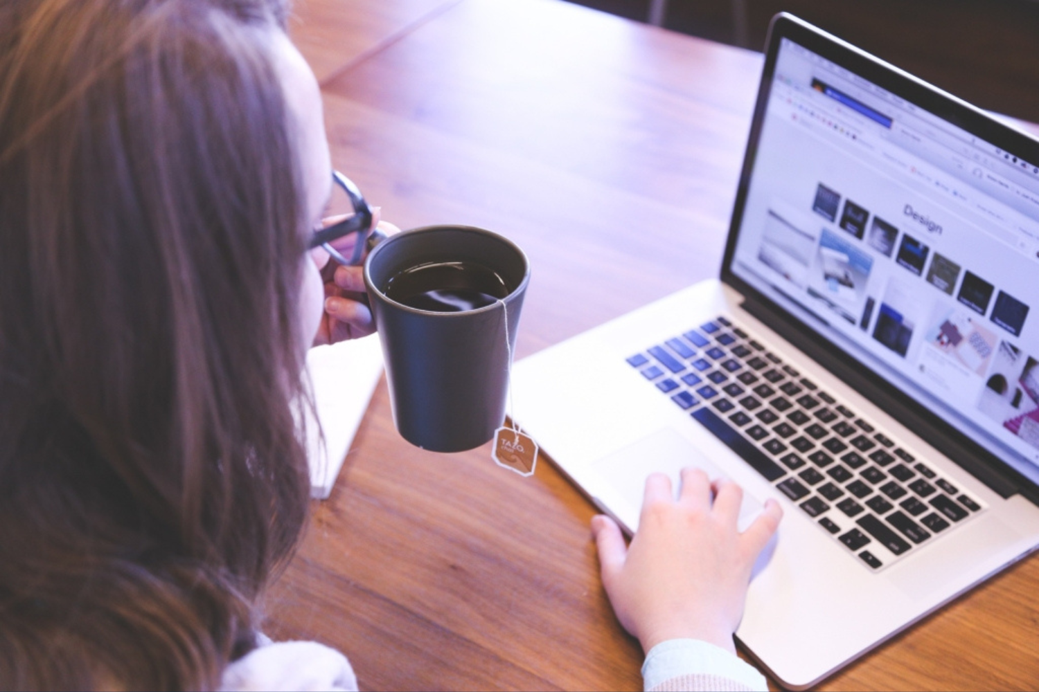 donna fotografata di spalle che sorseggia un tè da una tazza mentre scorre su un sito web con il suo pc appoggiato ad un tavolo in legno