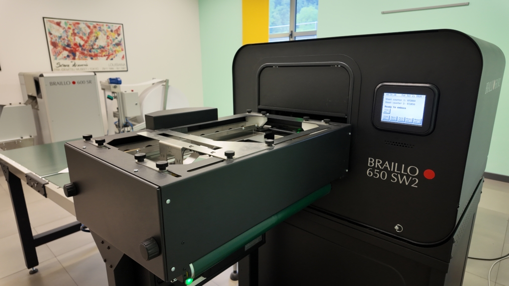 Stampante Braille industriale Braillo 650 SW2 con display digitale acceso, in una sala con pareti verdi e gialle; sullo sfondo un'altra macchina Braillo 600 SR.
