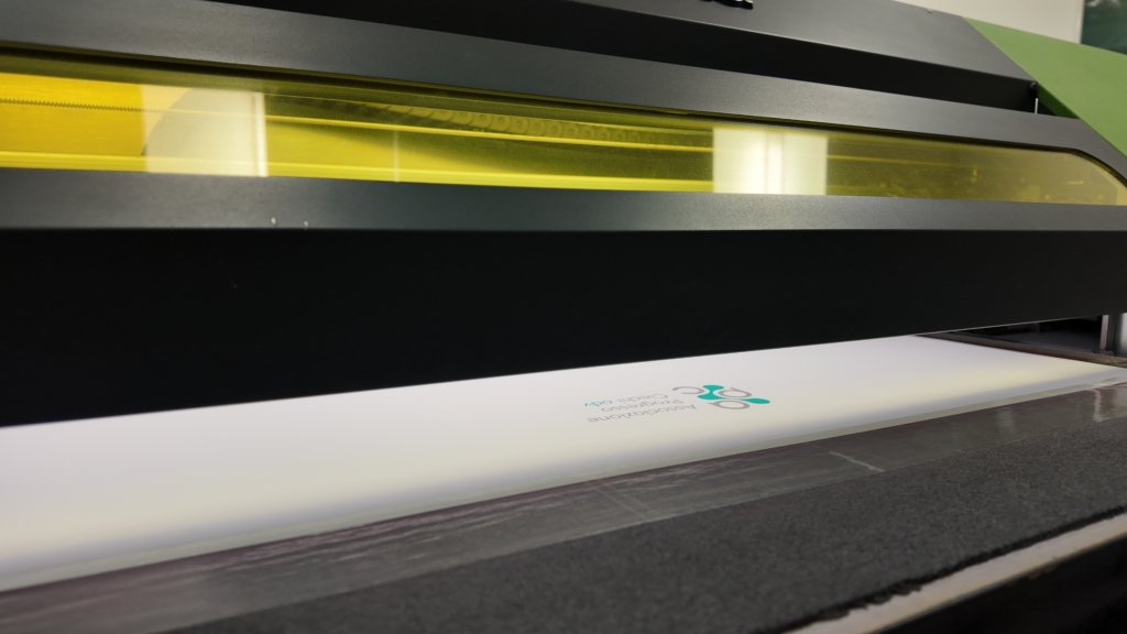 Primo piano del logo APC stampato su un pannello bianco, illuminato da luce UV blu all'interno di una stampante.