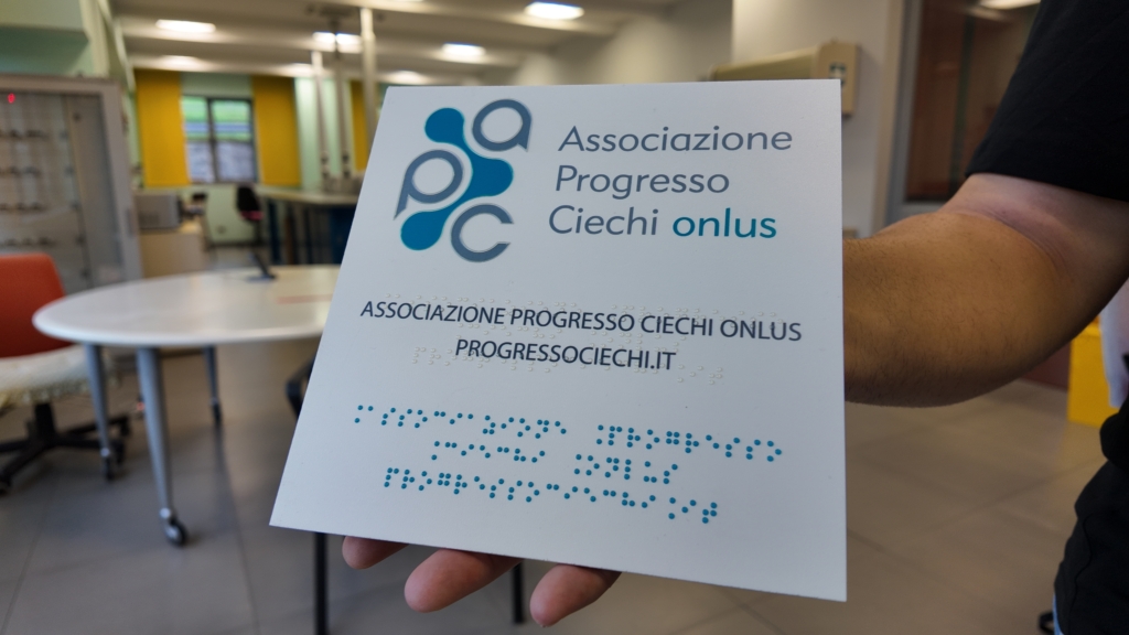 Una mano regge una targa bianca con il logo e il testo "Associazione Progresso Ciechi onlus" in caratteri stampati e in Braille, in un ambiente con arredi colorati sullo sfondo.