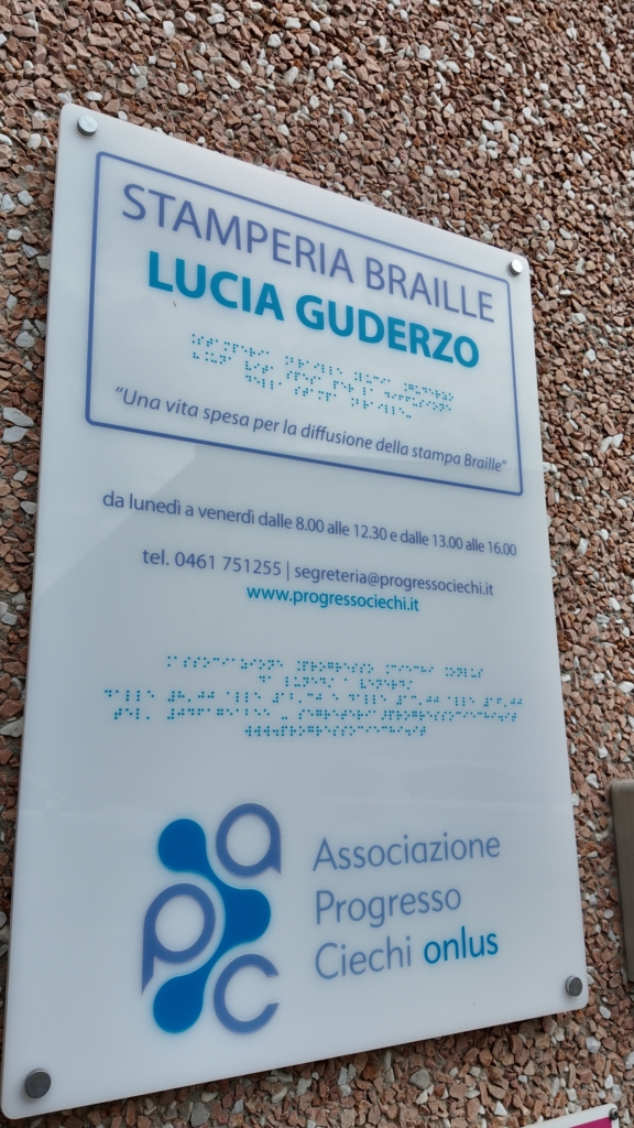 Targa esterna in plexiglass dedicata a "Stamperia Braille Lucia Guderzo", con testo anche in Braille e la frase "Una vita spesa per la diffusione della stampa Braille", fissata su una parete in pietra.