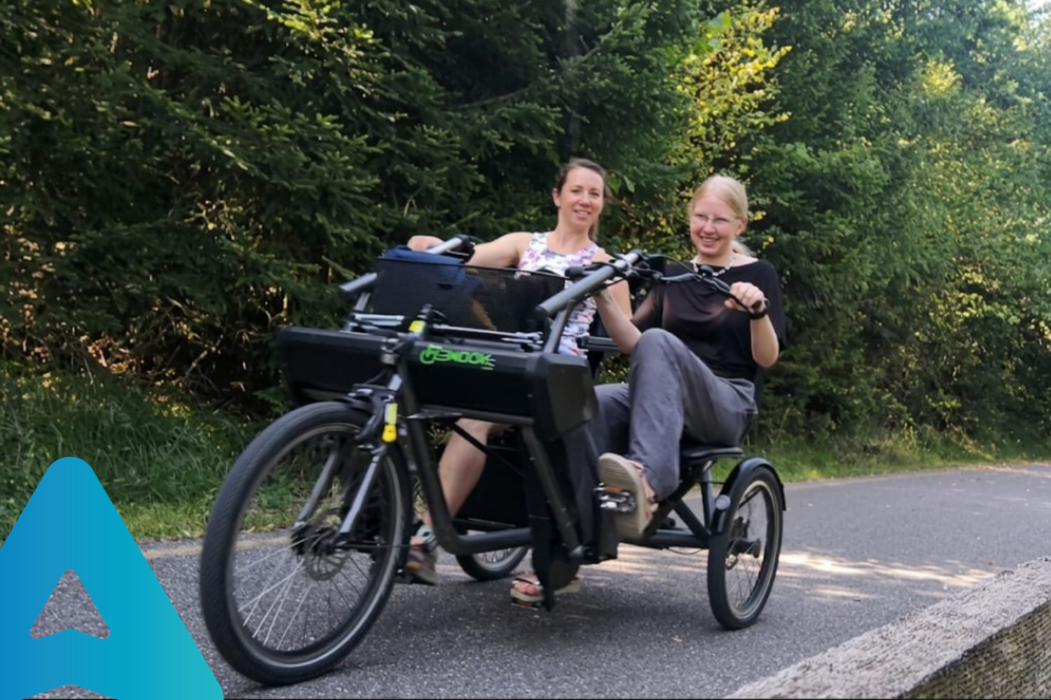 Due donne sorridenti su un handbike a tre ruote lungo una pista ciclabile immersa nel verde della Val di Fiemme, durante il tirocinio di accessibilità turistica di AbilNova.