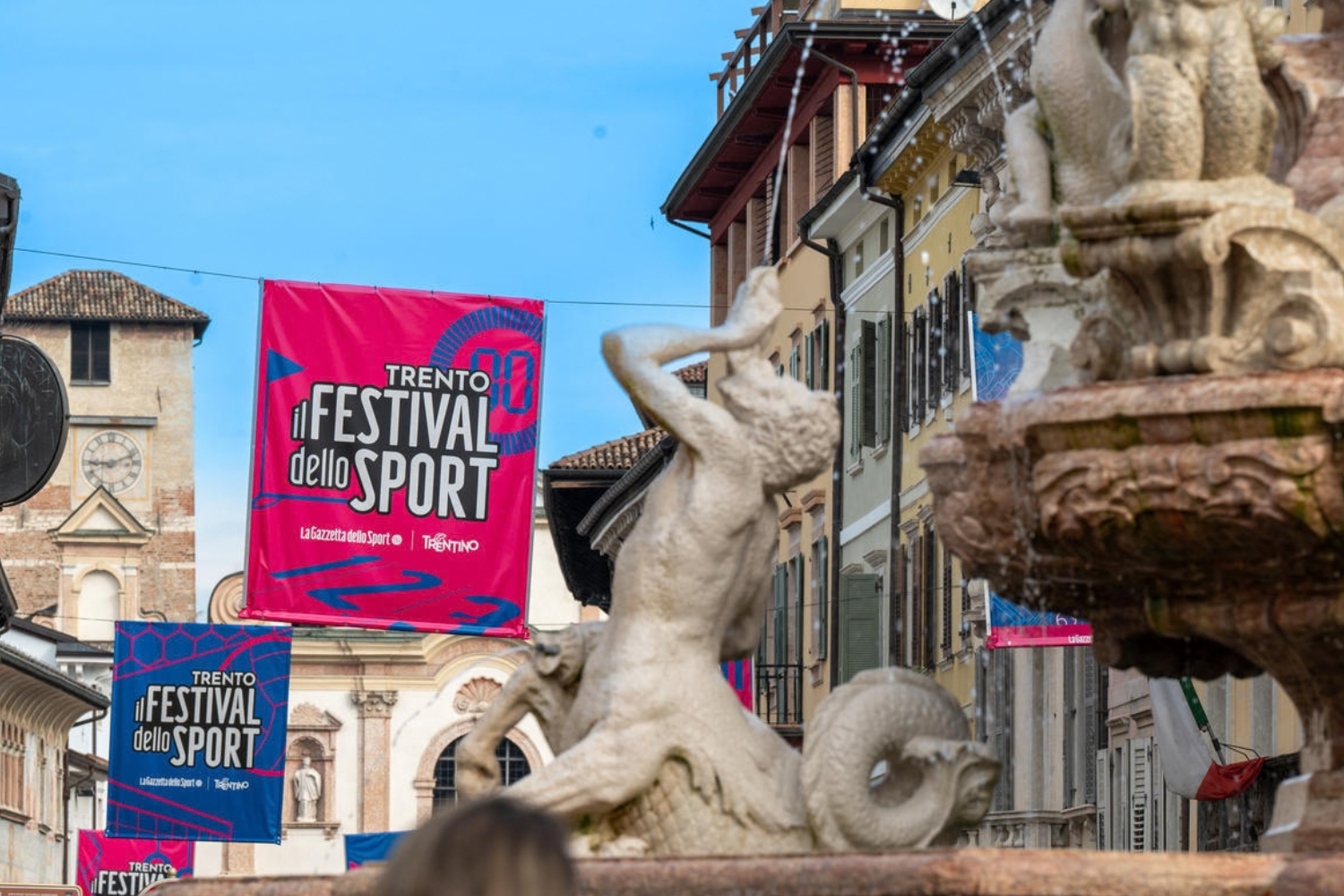 Bandiere rosa e blu del Festival dello Sport appese in una via del centro di Trento, con la fontana del Nettuno in primo piano.