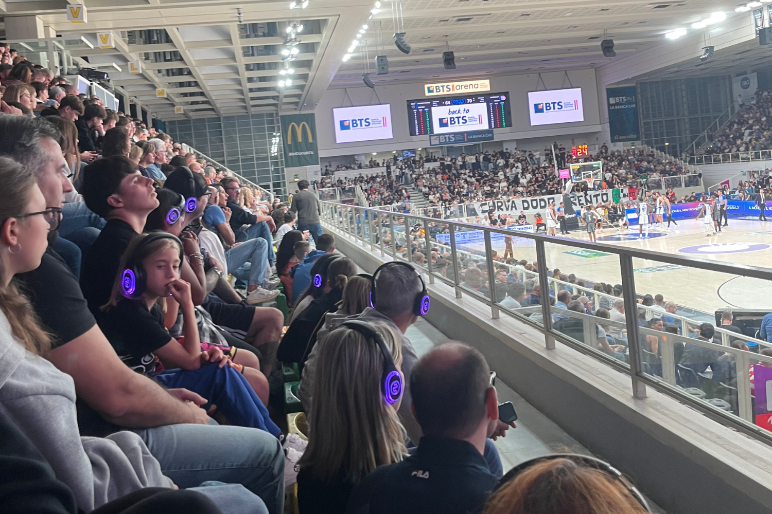 Persone di diverse età assistono a una partita di basket alla BTS Arena di Trento, alcune indossando cuffie per un’esperienza di ascolto dedicata, mentre sullo sfondo si vedono il campo da gioco e le tribune affollate.