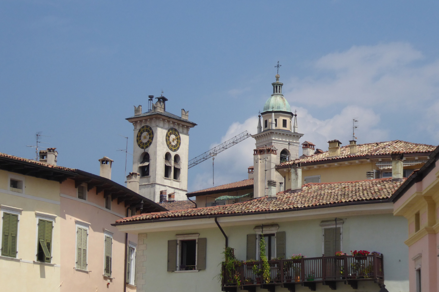 Veduta dei tetti del centro storico di Rovereto, con la Torre Civica e il campanile sullo sfondo, sotto un cielo azzurro con nuvole.